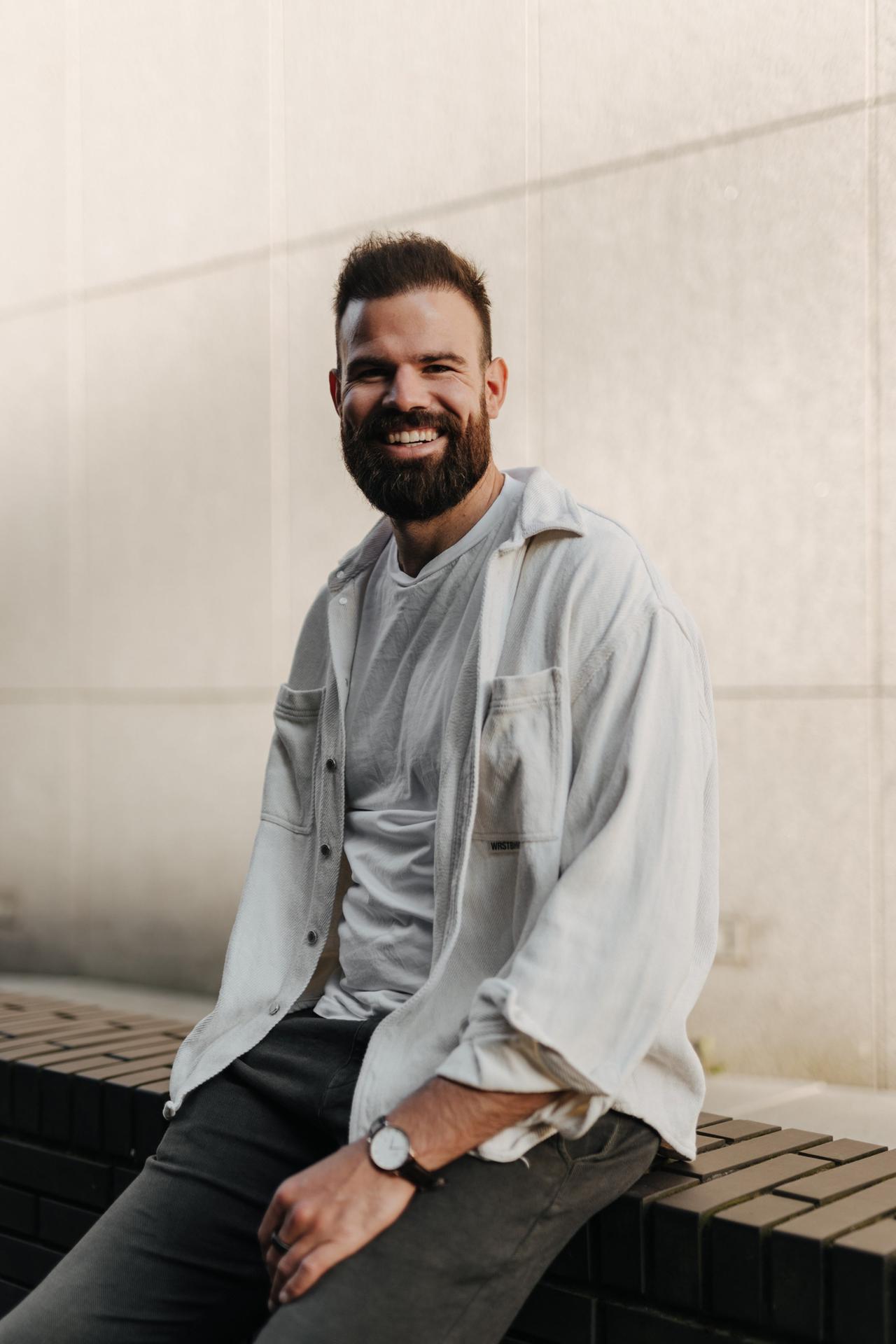Maximilian Planer sitzt auf einer Mauer und lächelt in die Kamera. Er trägt ein graues T-Shirt und einen grauen Pullover. Maximilian Planer sitzt auf einer Mauer und lächelt in die Kamera. Er trägt ein graues T-Shirt und einen grauen Pullover.