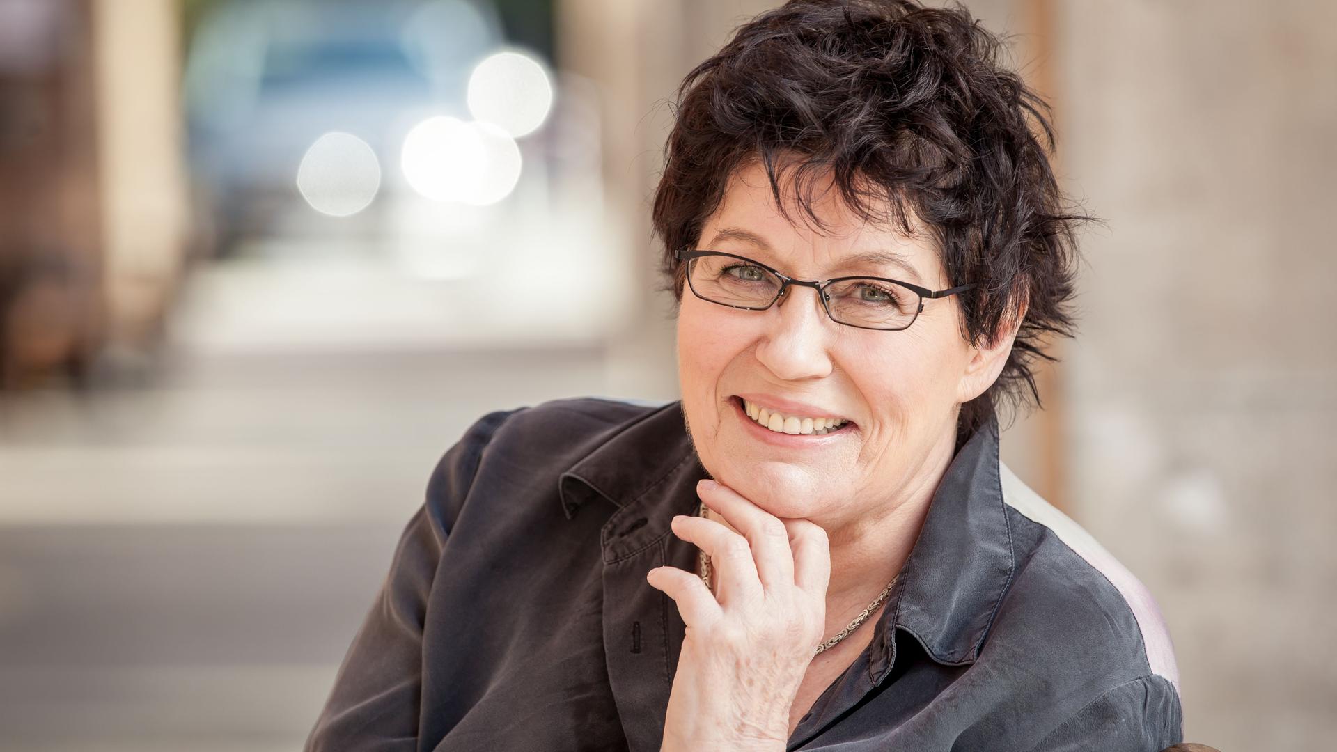 Uschi Brüning sitzt mit Brille und kurzen, dunklen Haaren vor der Kamera und lächelt, während sie ihr Kinn in ihre Hand stützt.