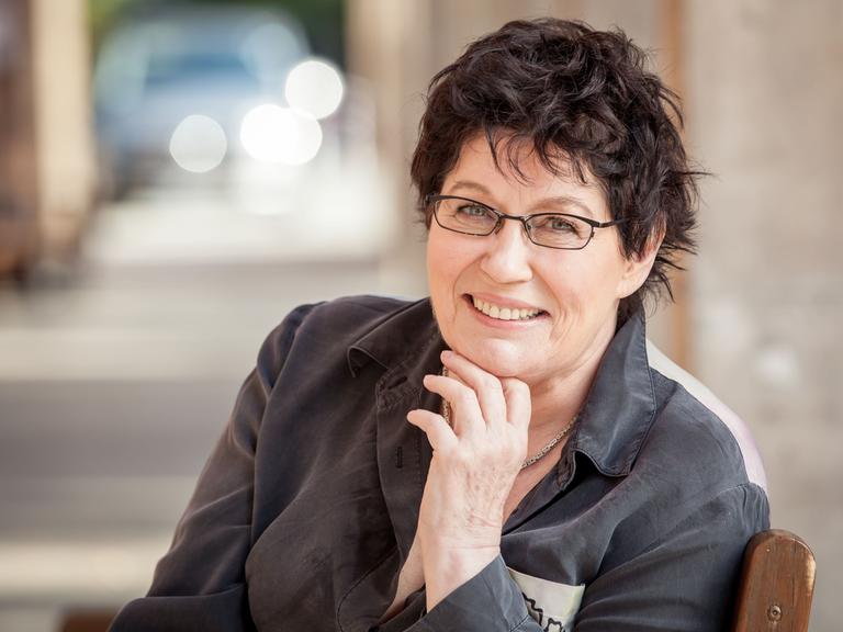 Uschi Brüning sitzt mit Brille und kurzen, dunklen Haaren vor der Kamera und lächelt, während sie ihr Kinn in ihre Hand stützt.