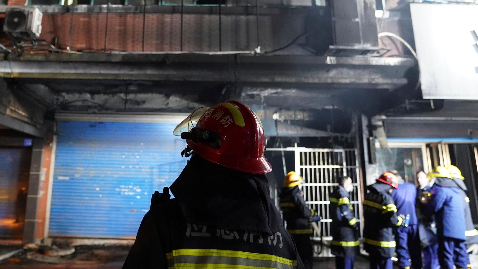 Man sieht von hinten mehrere Feuerwehrleute, die vor der teils schwarz verkohlten Gebäudefraont stehen. Man sieht von hinten mehrere Feuerwehrleute, die vor der teils schwarz verkohlten Gebäudefraont stehen.