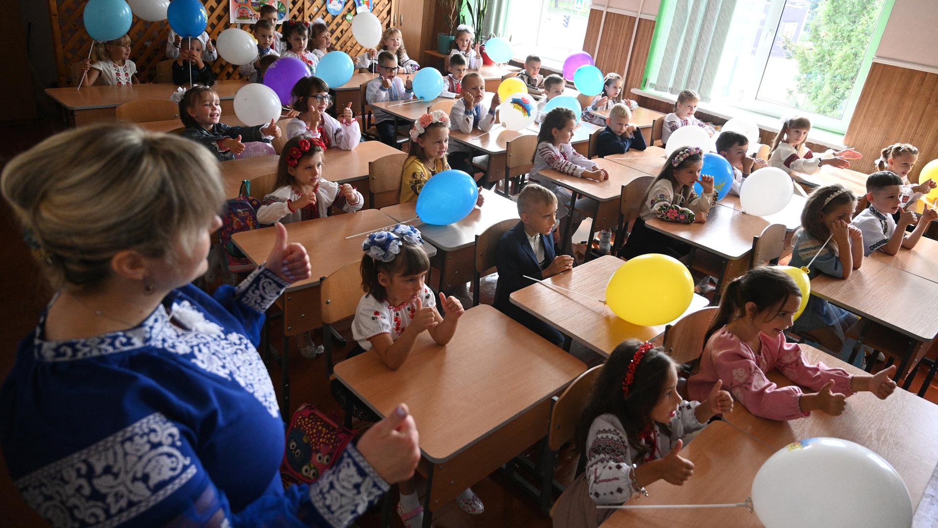 Schüler sitzen in einem Klassenzimmer und haben Lufballons in den Farben der Ukraine in den Händen.