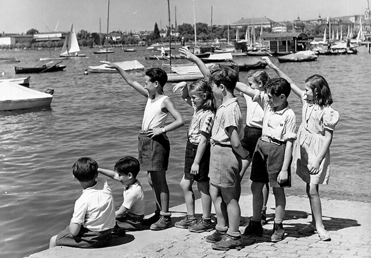 Acht Kinder stehen auf dem Schwarz.Weiß-Bild an einer Kaimauer, im Hintergrund sind Boote zu sehen, das Wasser ist ruhig,