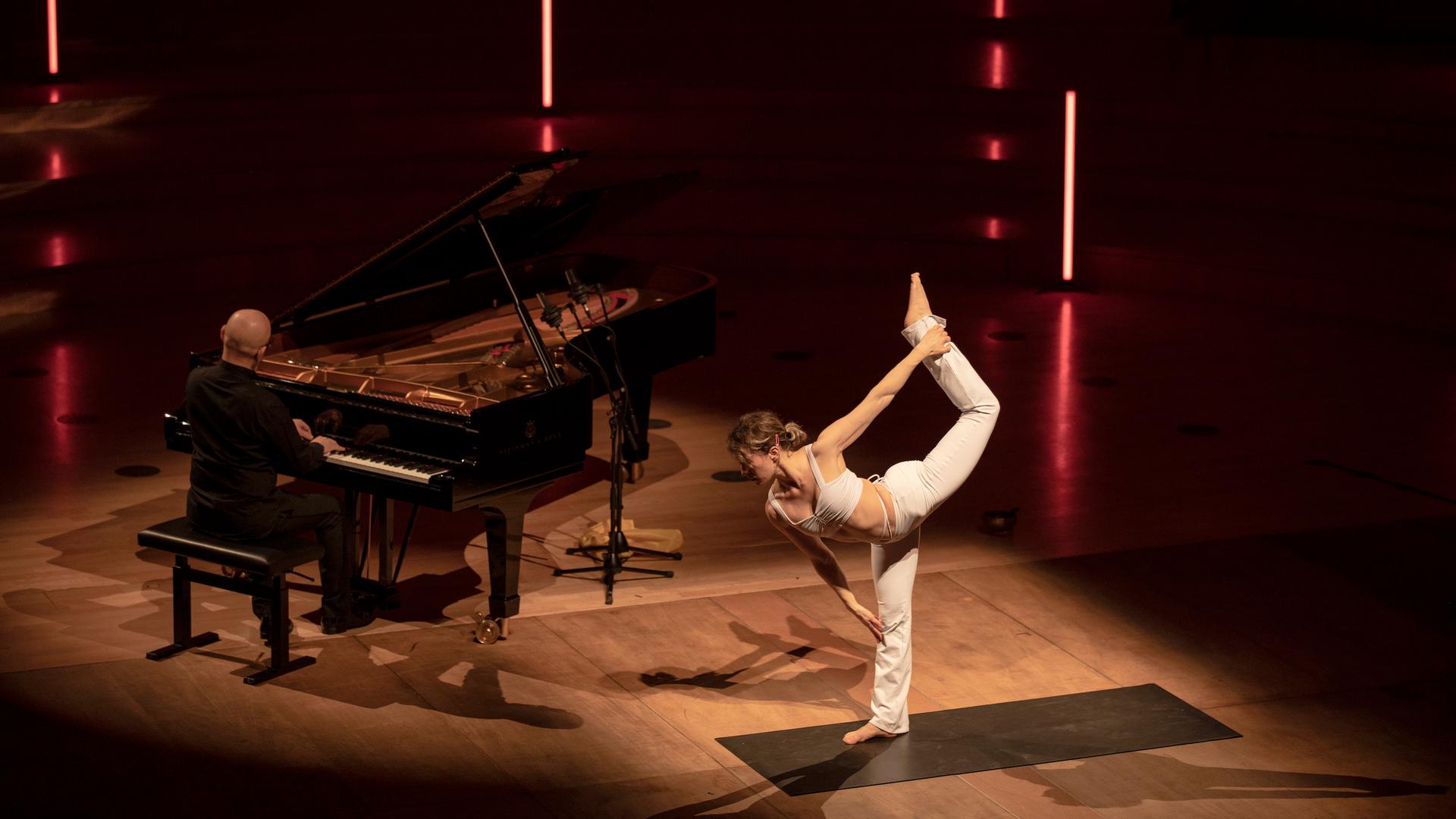 Anastasia Shevchenko nimmt auf der Bühne des Dresdner Kulturpalastes eine Yoga-Pose ein, links von ihr sitzt Ohad Ben-Ari an einem Flügel. Shevchenko trägt weiße Yoga-Kleidung.