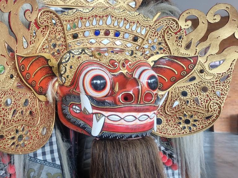 Blick auf eine großformatige Maske mit großen schwarzen Augen, großen Ohren und roter Haut, die mit einer aufwendigen Krone geziert ist.