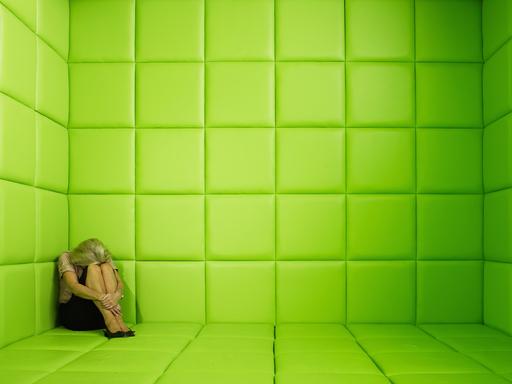 Eine Frau sitzt in der Ecke einer grün verkleideten Gummizelle und verbirgt den Kopf in ihren Armen.