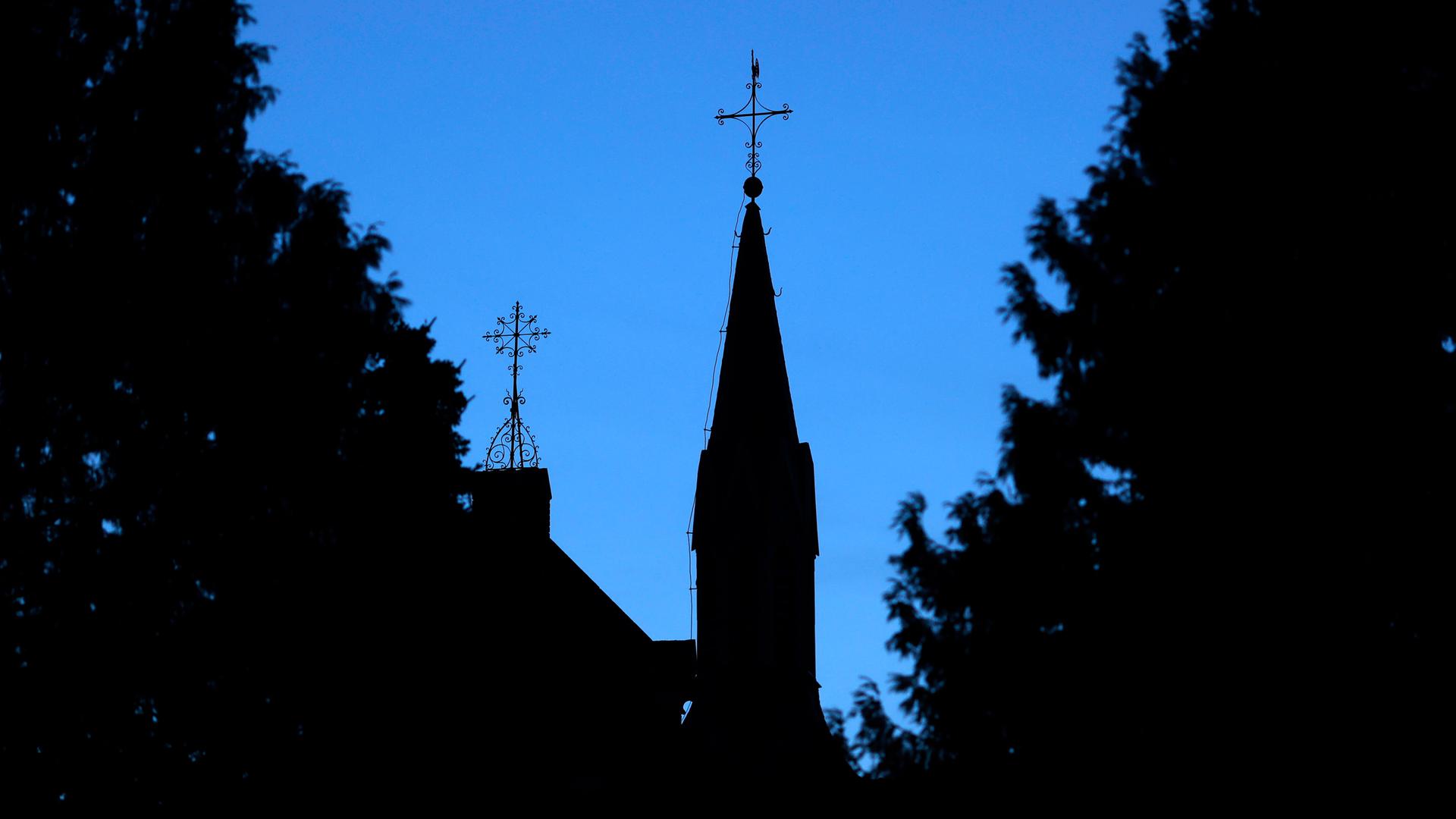 Ein Kreuz mit Wetterhahn steht auf dem Dach einer Kirche vor einem dunkelblauen Abendhimmel. 