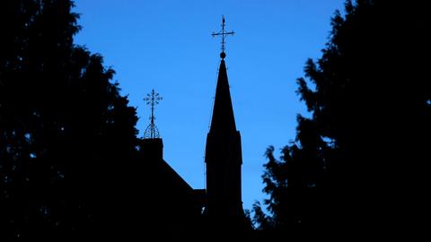 Ein Kreuz mit Wetterhahn steht auf dem Dach einer Kirche vor einem dunkelblauen Abendhimmel. 