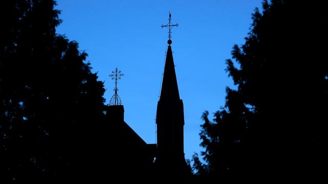 Ein Kreuz mit Wetterhahn steht auf dem Dach einer Kirche vor einem dunkelblauen Abendhimmel. 