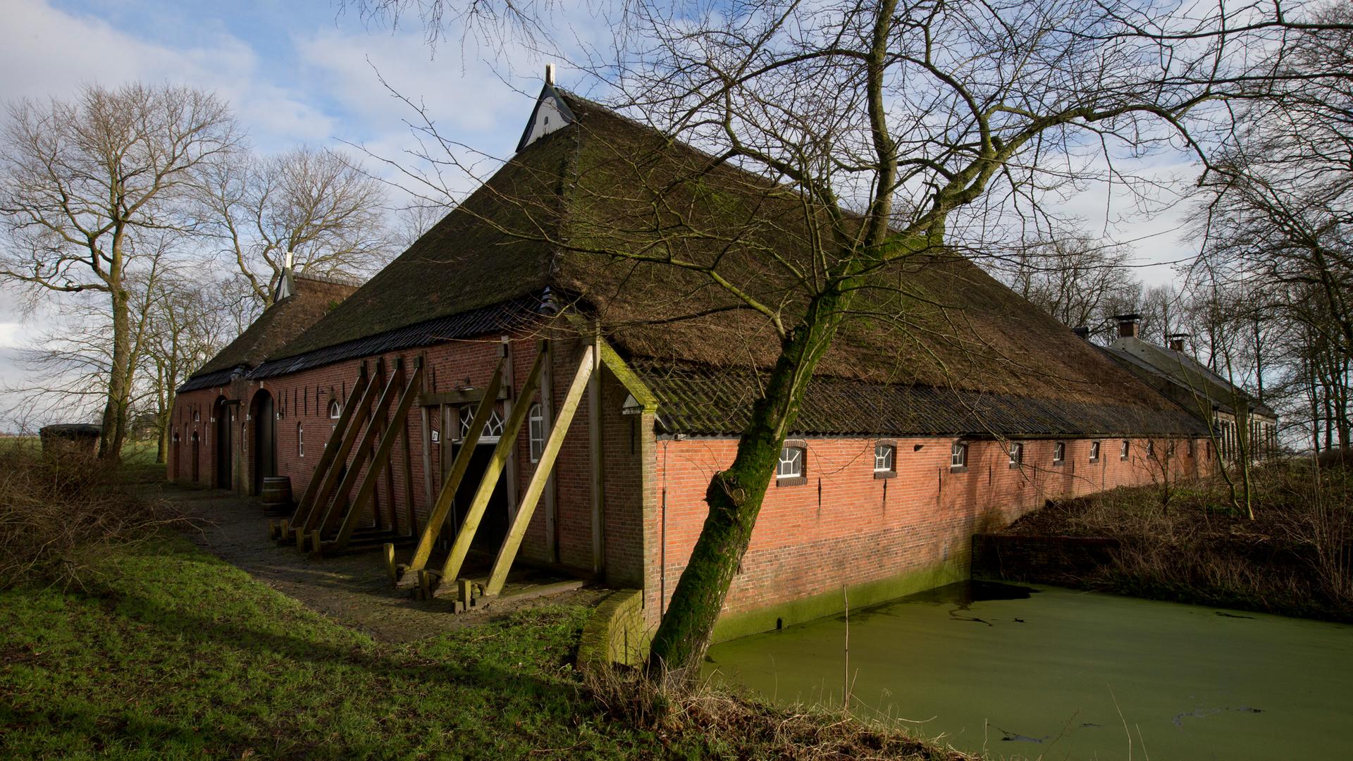 Zu sehen ist ein traditionelles Backsteinhaus in ländlicher Umgebung. Das Gebäude wird von mehreren schräg angebrachten Stützbalken stabilisiert, die an der Außenwand lehnen.