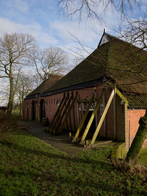 Zu sehen ist ein traditionelles Backsteinhaus in ländlicher Umgebung. Das Gebäude wird von mehreren schräg angebrachten Stützbalken stabilisiert, die an der Außenwand lehnen.