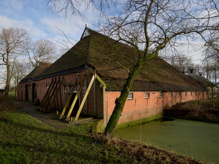 Zu sehen ist ein traditionelles Backsteinhaus in ländlicher Umgebung. Das Gebäude wird von mehreren schräg angebrachten Stützbalken stabilisiert, die an der Außenwand lehnen.