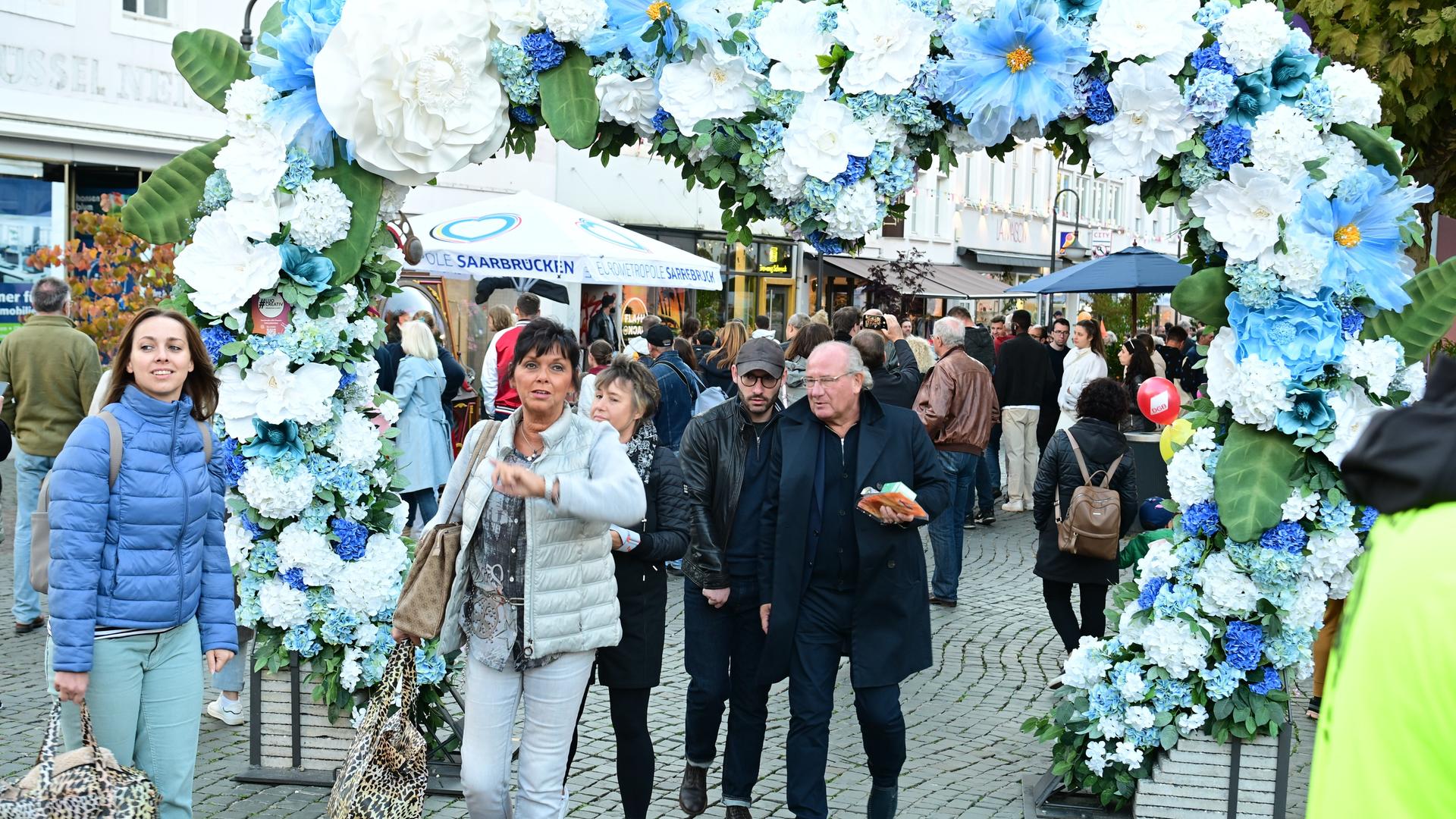 Bürgerinnen und Bürger laufen durch die Saarbrücker Innenstadt, im Vordergrund eine Blumengirlande