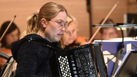 Eine junge Frau mit blondem Pferdeschwanz steht zwischen einem Orchester und einem Notenständer und spielt konzentriert Akkordeon.