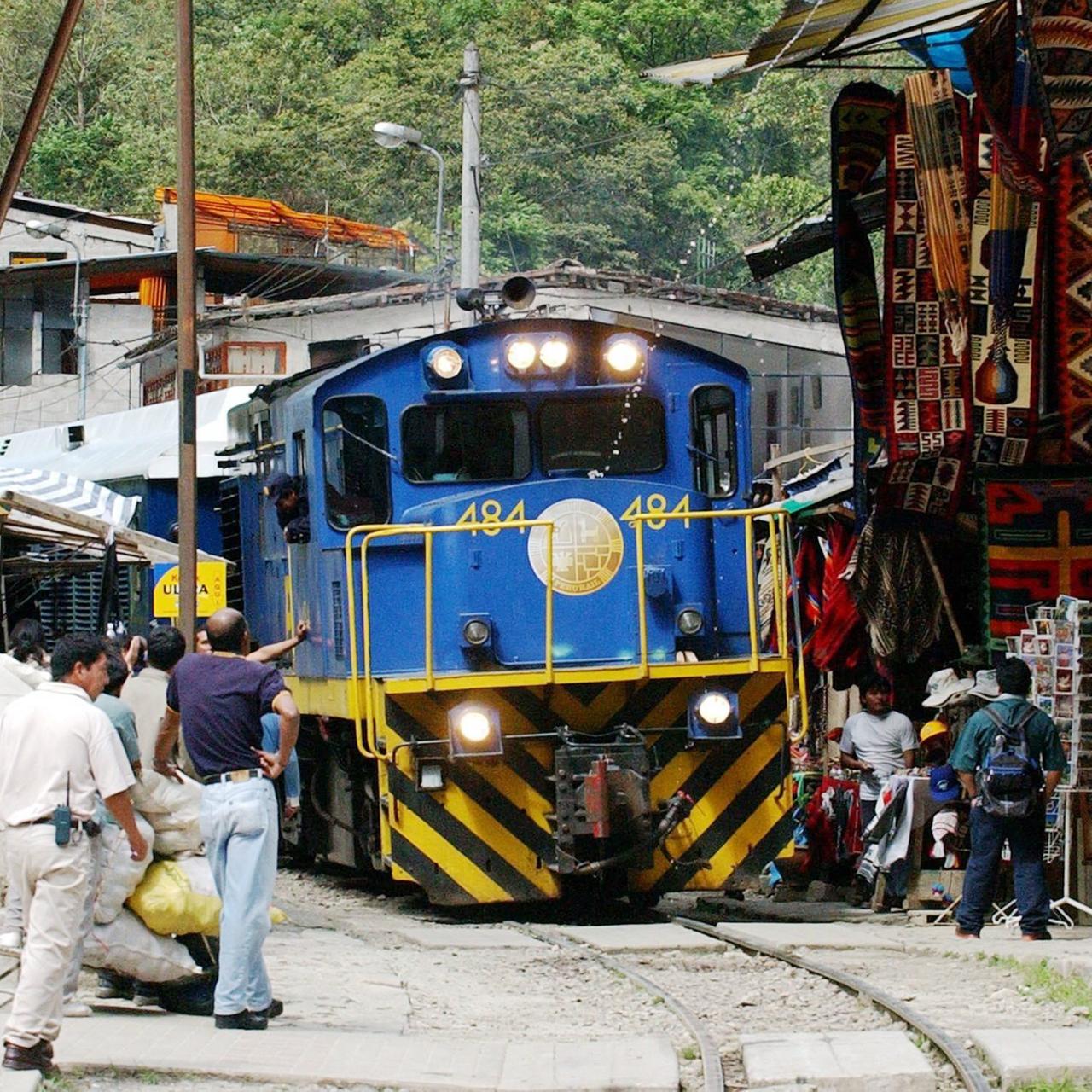 Blick von vorne auf den blauen Triebwagen mit gelben Streifen. Rechts Andenkenstände, links mehrere Personen neben den Gleisen.