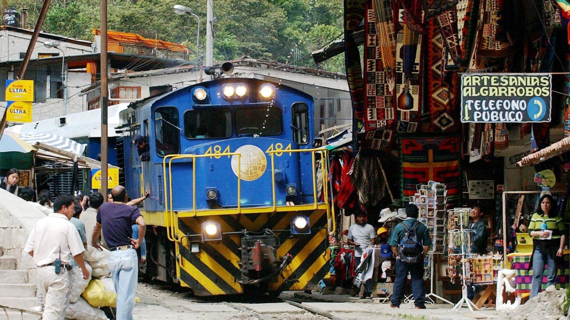 Blick von vorne auf den blauen Triebwagen mit gelben Streifen. Rechts Andenkenstände, links mehrere Personen neben den Gleisen. Blick von vorne auf den blauen Triebwagen mit gelben Streifen. Rechts Andenkenstände, links mehrere Personen neben den Gleisen.