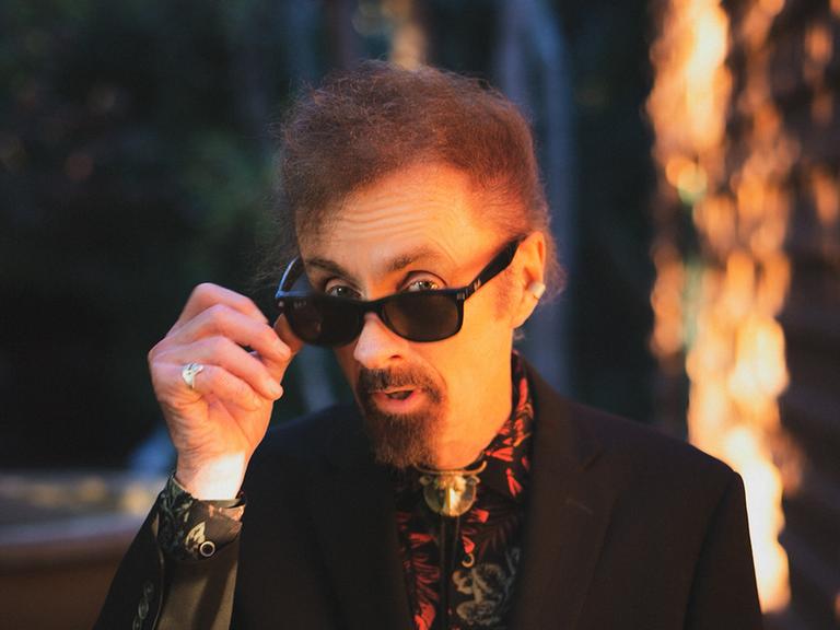 Der Schriftsteller T.C Boyle guckt im schwarzen Jacket und sehr bunten Hemd über seine Sonnenbrille hinweg. Im Hintergrund sind unscharf Bäume zu erahnen.