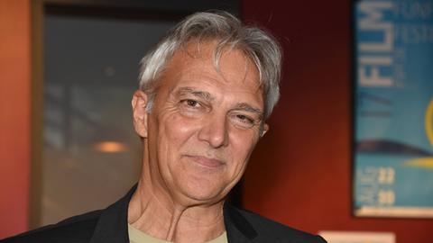 Matthias Helwig guckt grinsend in die Kamera. Er trägt ein beigefarbenes T-Shirt und eine schwarze Jacke.