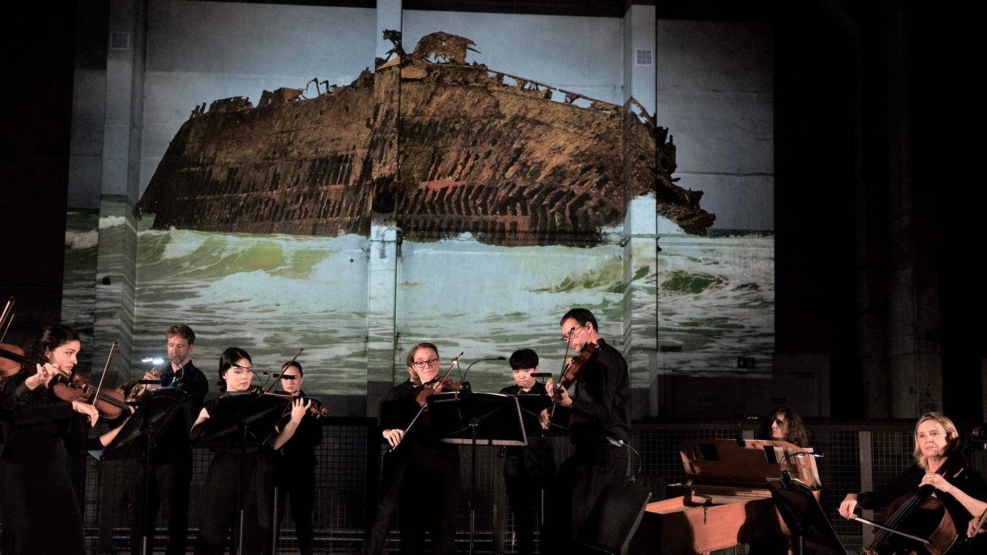 Ein Musikensemble mit Streichern, einer Cembalistin, einem Hornisten und einem Oboisten spielt in schwarzer Konzertkleidung auf einer Bühne. Im Hintergrund wird ein Foto an die Wand projiziert, das ein auf Grund gelaufenes, zur Seite geneigtes Schiffswrack zeigt.