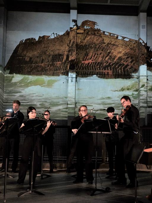 Ein Musikensemble mit Streichern, einer Cembalistin, einem Hornisten und einem Oboisten spielt in schwarzer Konzertkleidung auf einer Bühne. Im Hintergrund wird ein Foto an die Wand projiziert, das ein auf Grund gelaufenes, zur Seite geneigtes Schiffswrack zeigt.