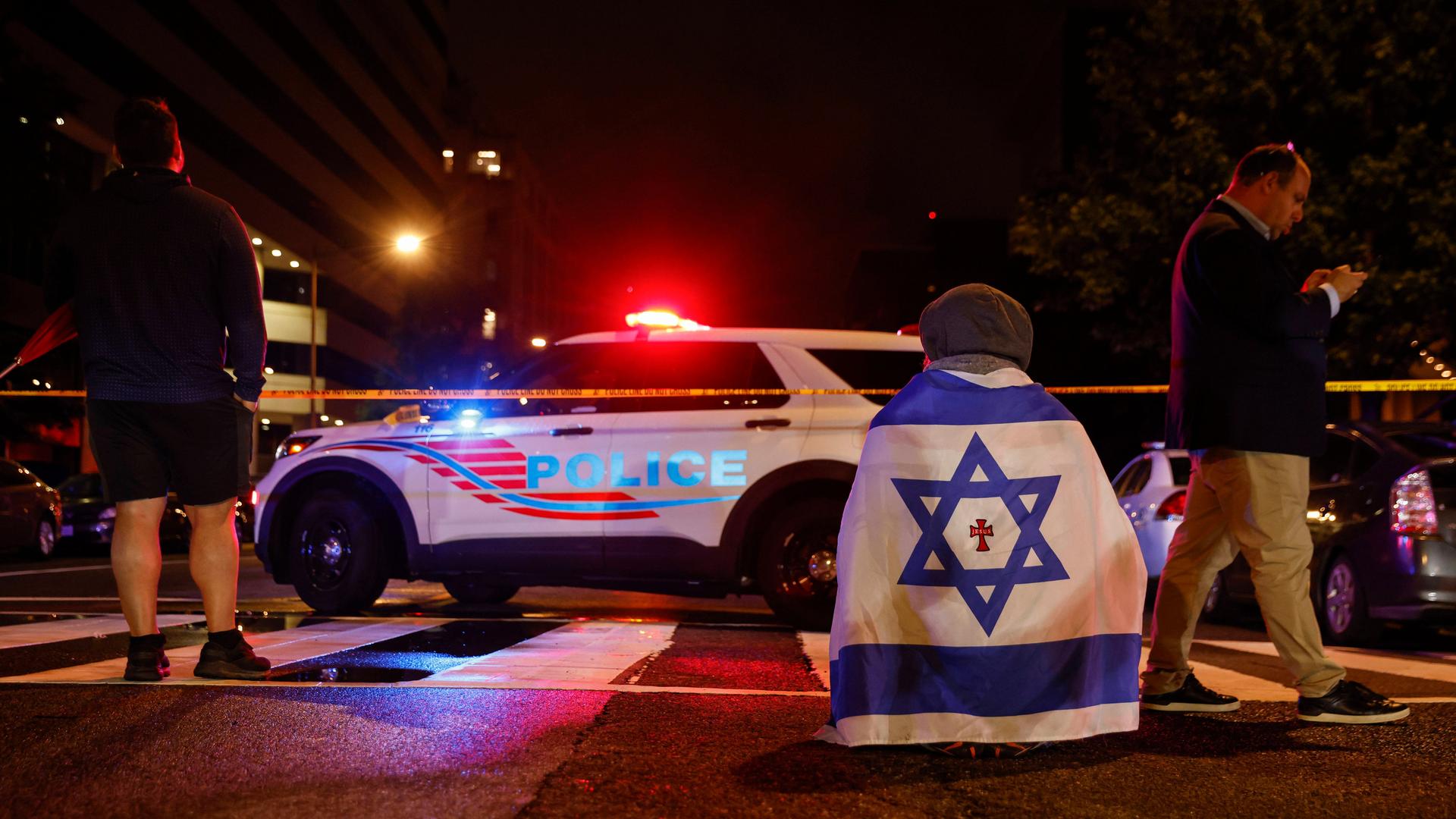 Ein Polizeiauto mit Blinklicht steht im Dunkeln amStraßenrand. Davor sitzt eine in eine israelische Flagge eingehüllte Person auf dem Boden, man sieht sie von hinten. 