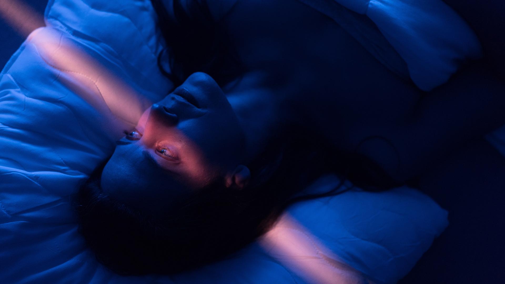 Eine Frau mit erschöpftem Gesichtsausdruck liegt in einem abgedunkelten Zimmer im Bett, ihre Augen sind geöffnet, ein leicht rosafarbener Lichtstrahl läuft vertikal durchs Bild. Eine Frau mit erschöpftem Gesichtsausdruck liegt in einem abgedunkelten Zimmer im Bett, ihre Augen sind geöffnet, ein leicht rosafarbener Lichtstrahl läuft vertikal durchs Bild.