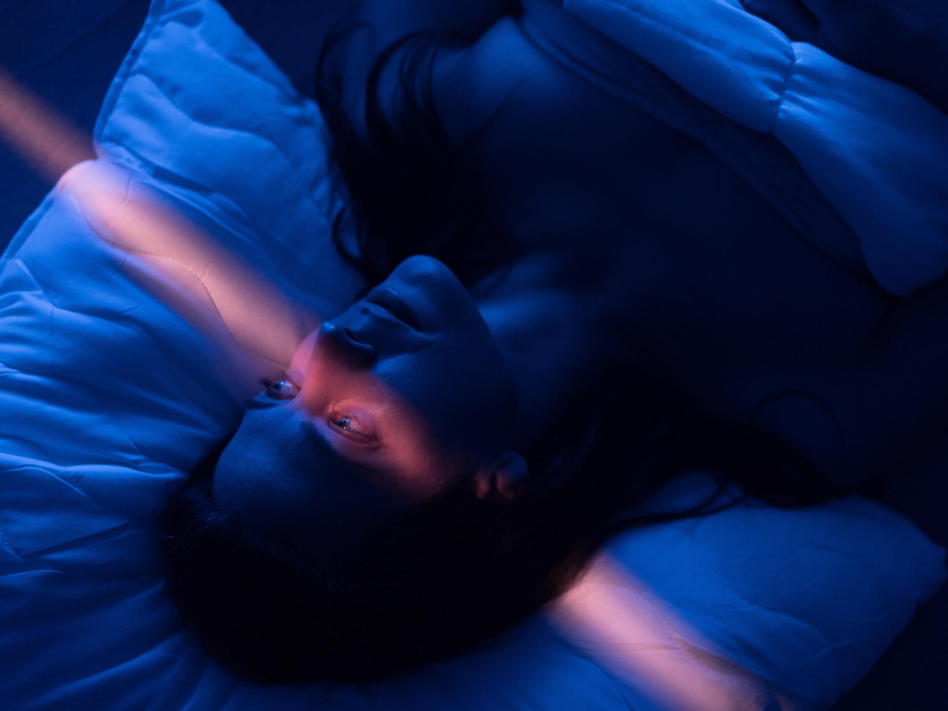 Eine Frau mit erschöpftem Gesichtsausdruck liegt in einem abgedunkelten Zimmer im Bett, ihre Augen sind geöffnet, ein leicht rosafarbener Lichtstrahl läuft vertikal durchs Bild.