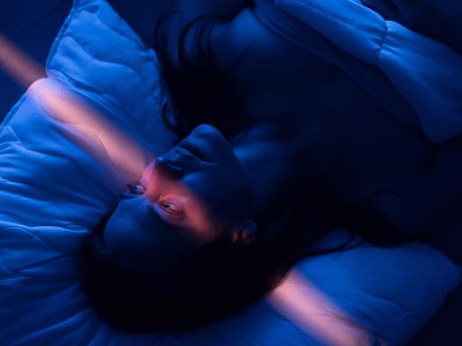 Eine Frau mit erschöpftem Gesichtsausdruck liegt in einem abgedunkelten Zimmer im Bett, ihre Augen sind geöffnet, ein leicht rosafarbener Lichtstrahl läuft vertikal durchs Bild.
