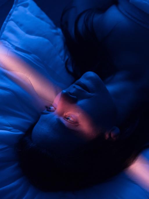 Eine Frau mit erschöpftem Gesichtsausdruck liegt in einem abgedunkelten Zimmer im Bett, ihre Augen sind geöffnet, ein leicht rosafarbener Lichtstrahl läuft vertikal durchs Bild. Eine Frau mit erschöpftem Gesichtsausdruck liegt in einem abgedunkelten Zimmer im Bett, ihre Augen sind geöffnet, ein leicht rosafarbener Lichtstrahl läuft vertikal durchs Bild.