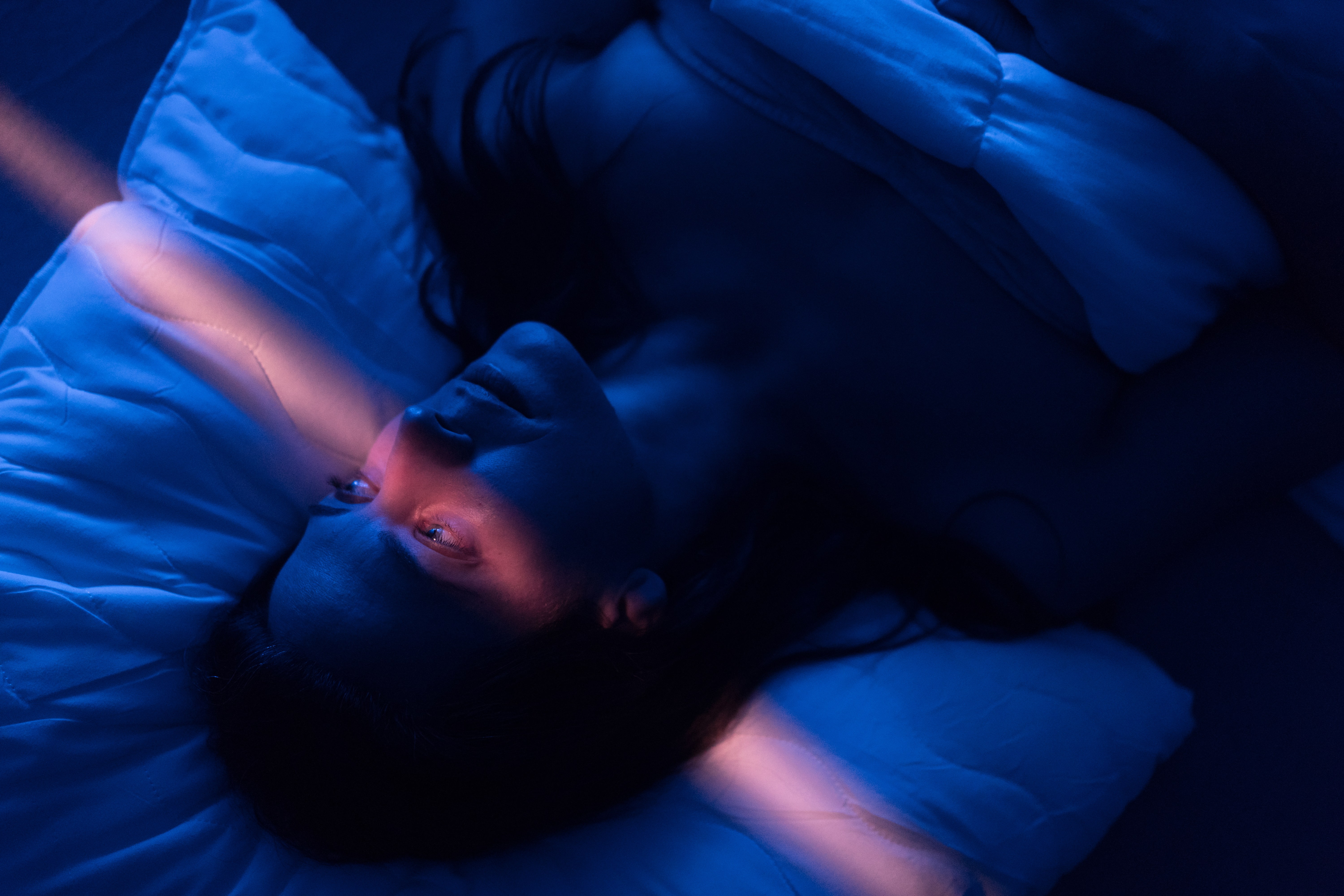 Eine Frau mit erschöpftem Gesichtsausdruck liegt in einem abgedunkelten Zimmer im Bett, ihre Augen sind geöffnet, ein leicht rosafarbener Lichtstrahl läuft vertikal durchs Bild. Eine Frau mit erschöpftem Gesichtsausdruck liegt in einem abgedunkelten Zimmer im Bett, ihre Augen sind geöffnet, ein leicht rosafarbener Lichtstrahl läuft vertikal durchs Bild.