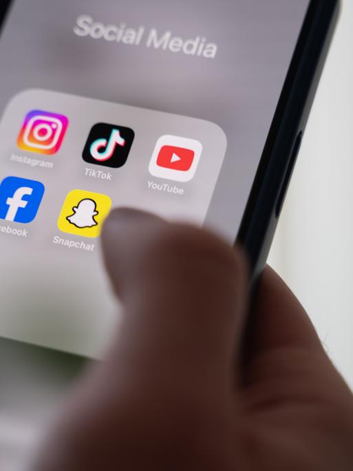 Eine Frau hält ein Smartphone, auf dessen Display verschiedene Social Media Apps angezeigt werden. 
