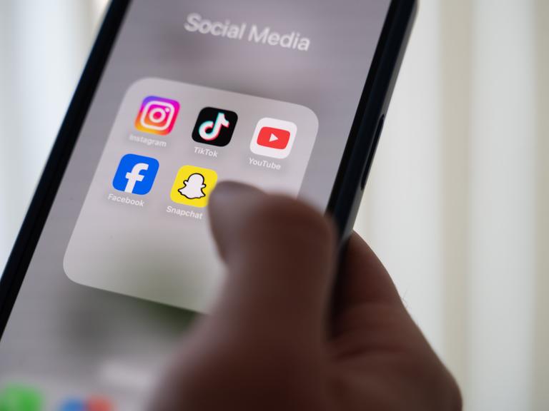 Eine Frau hält ein Smartphone, auf dessen Display verschiedene Social Media Apps angezeigt werden. 