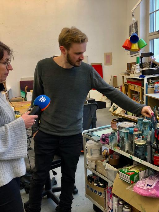 Eine dunkelhaarige Frau mit Mikrofon in der Hand und ein rotblonder junger Mann vor einem Atelierwagen mit vielen Malutensilien