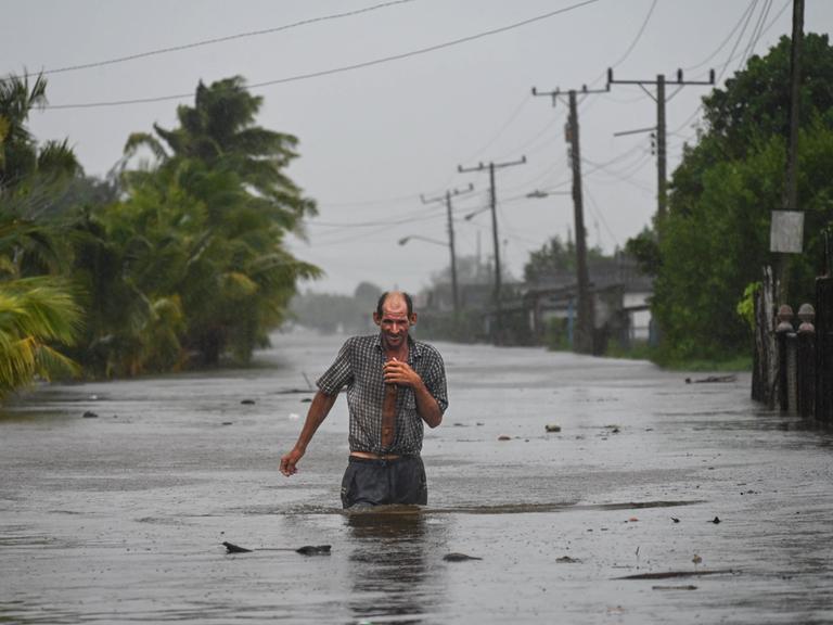 Ein Mann watet durch eine geflutete Straße, das Wasser reicht ihm bis zur Hüfte.