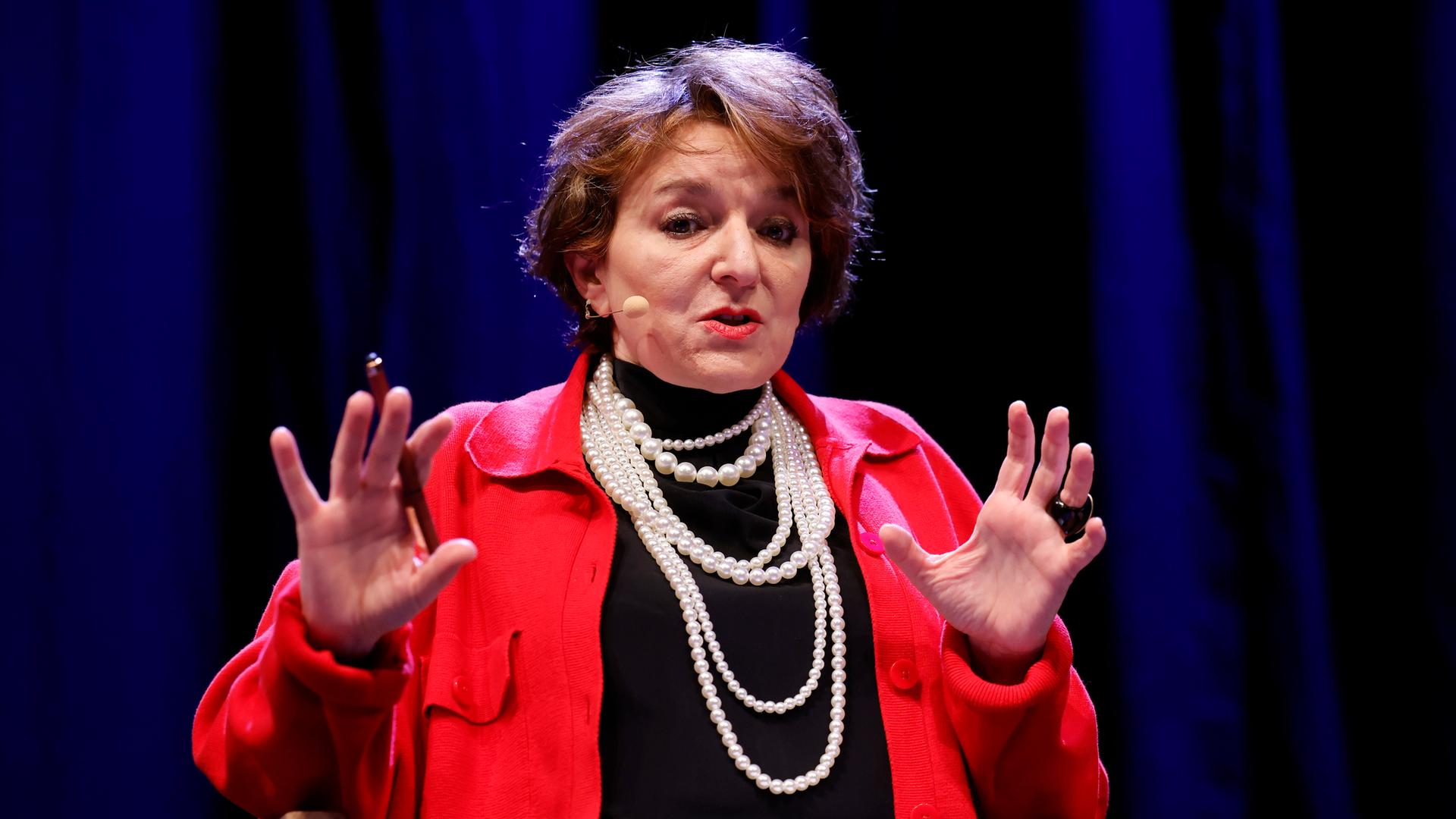 Die Soziologin Eva Illouz sitzt auf einer Podiumsbühne. Sie trägt eine rote Jacke, schwarzen Rollkragenpullover und mehrere helle Perlenketten um den Hals. Im Hintergrund: Ein dunkelblauer Vorhang.