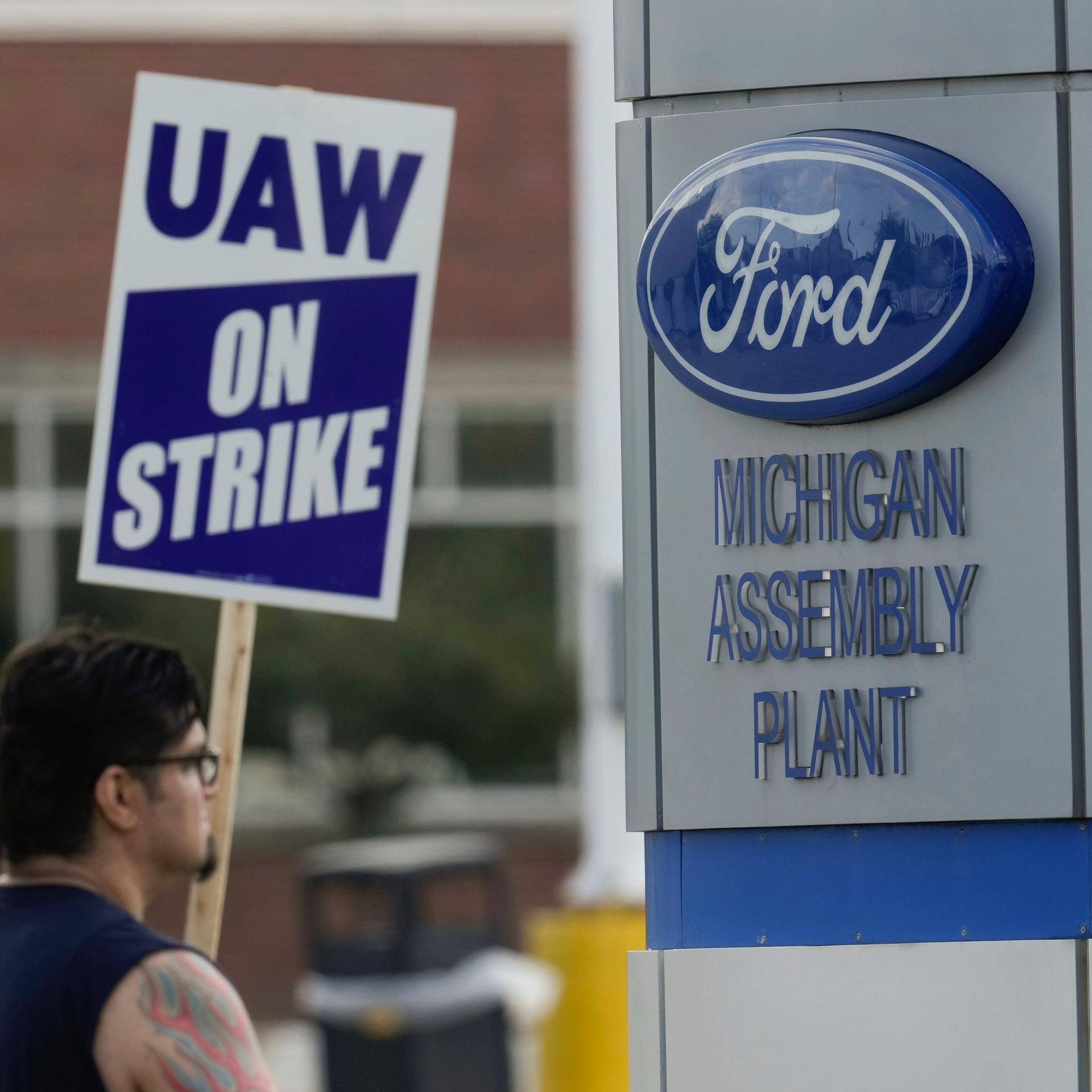 Ein Streikender mit einem Schild der Gewerkschaft UAW neben einem Logo des Autobauers Ford. Ein Streikender mit einem Schild der Gewerkschaft UAW neben einem Logo des Autobauers Ford.