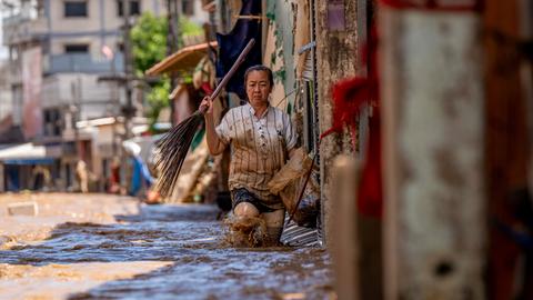 Eine Frau läuft auf einer Straße in Mae Sai in Nordthailand knietief im Hochwasser und trägt in einer Hand einen Besen.
