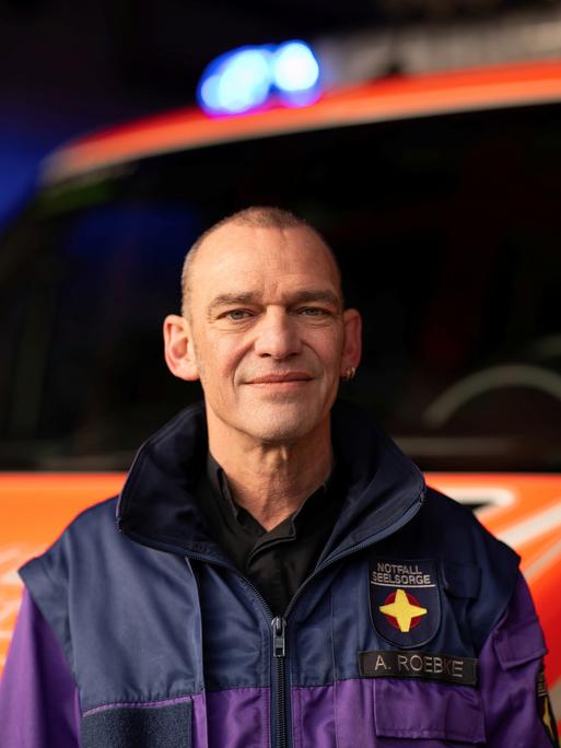 Albi Roebke trägt eine Jacke der Notfallseelsorge und steht vor einem Rettungswagen.