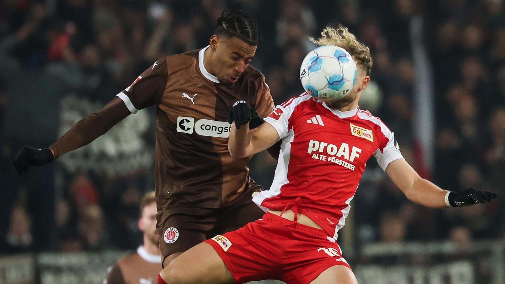 Die beiden springen zum Kopfball hoch. Guliavogui trägt ein braunes Trikot; Hollerbach ein weiß-rotes. Sein Kopf ist vom Ball verdeckt. 