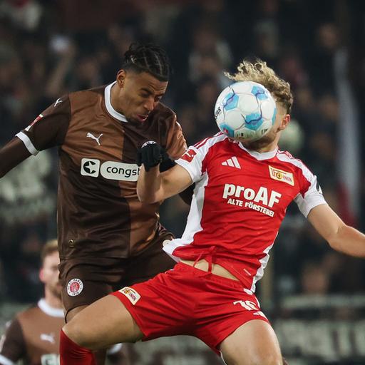 Die beiden springen zum Kopfball hoch. Guliavogui trägt ein braunes Trikot; Hollerbach ein weiß-rotes. Sein Kopf ist vom Ball verdeckt. 