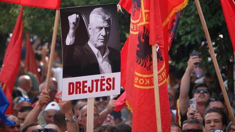 Zu sehen ist eine Menschenmenge. Darüber wehen rote Kosovo-Fahnen sowie ein Plakat, das Hashim Thaci abbildet. Darunter steht „drejtësi“ – das kosovarische Wort für „Gerechtigkeit“.