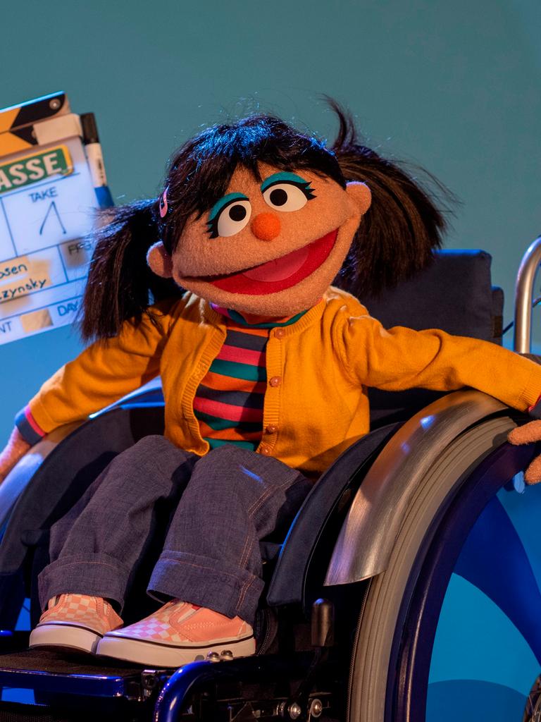 Eine Puppe aus der Sesamstraße, ein Mädchen mit Zöpfen, sitzt im Rollstuhl. 