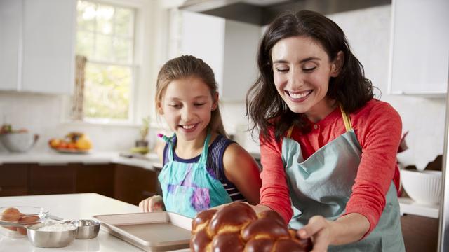 Eine Frau mit braunen Haaren und mit Schürze steht in der Küche. Sie hält einen Hefezopf in der Hand. Links neben hier steht ihre Tochter. Eine Frau mit braunen Haaren und mit Schürze steht in der Küche. Sie hält einen Hefezopf in der Hand. Links neben hier steht ihre Tochter.