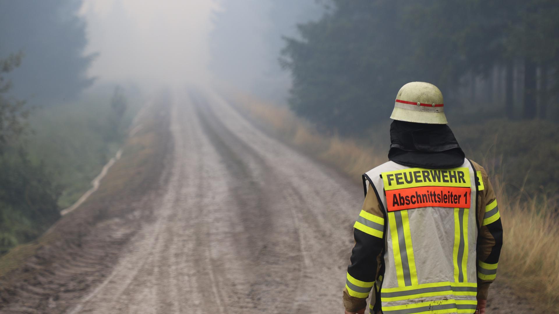 Ein Feuerwehrmann in Rückansicht steht auf einem Weg in einem verqualmten Waldstück im Hohen Venn.