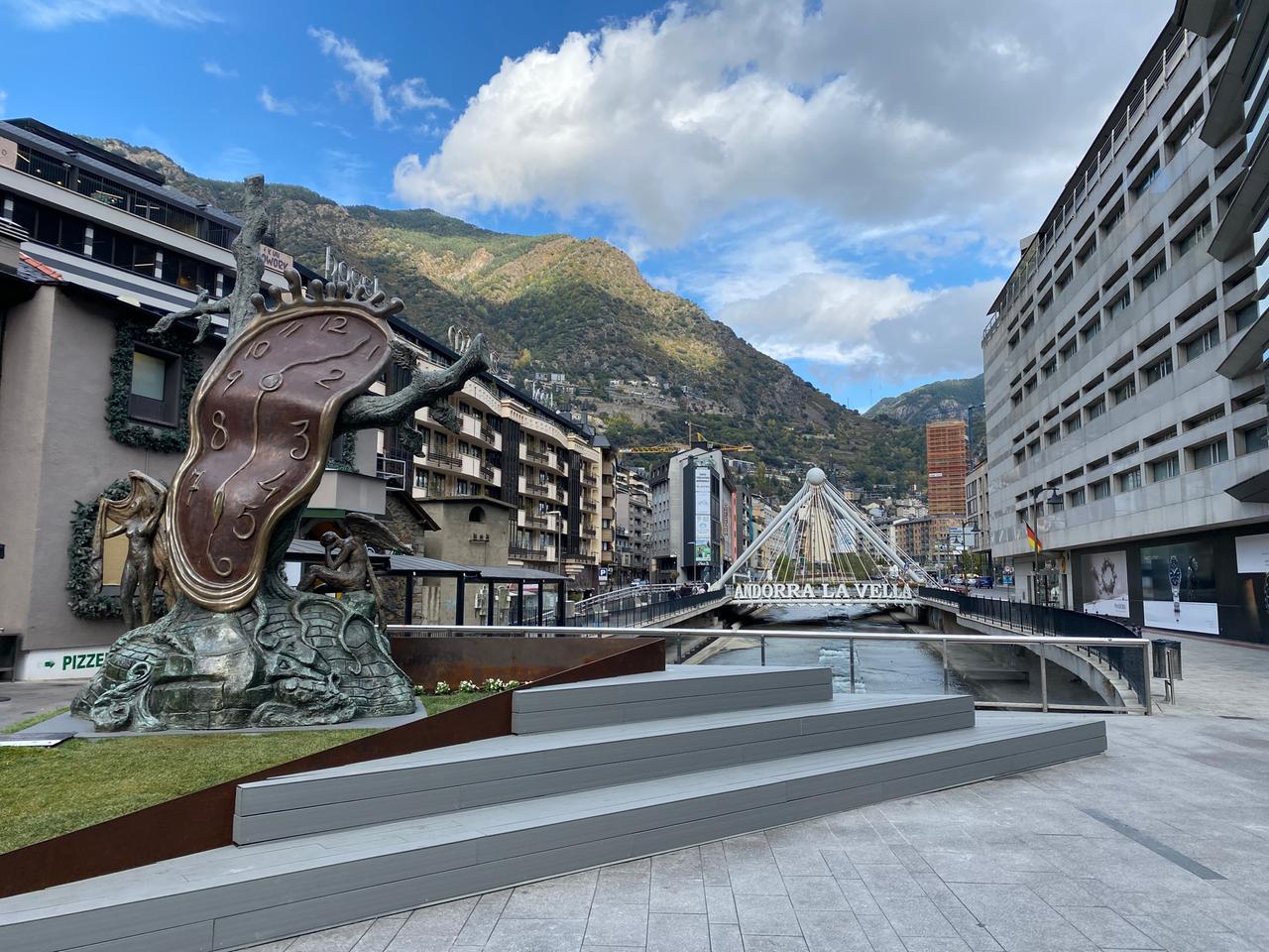 Das bekannteste Fotomotiv von Andorra - die Fußgängerzone inmitten der Berge, im Vordergrund eine große schiefe Uhr und über dem Fluß der Schriftzug "Andorra La Vella".