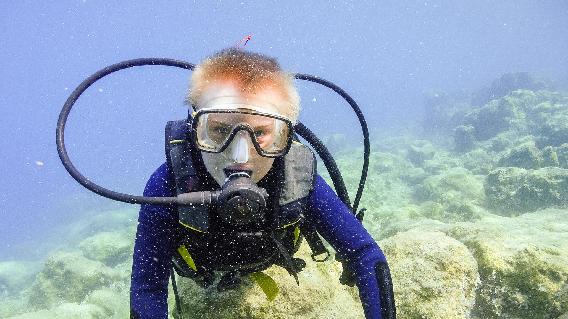 Ein Junge in voeller tauchermontur unter Wasser schaut in die Kamera