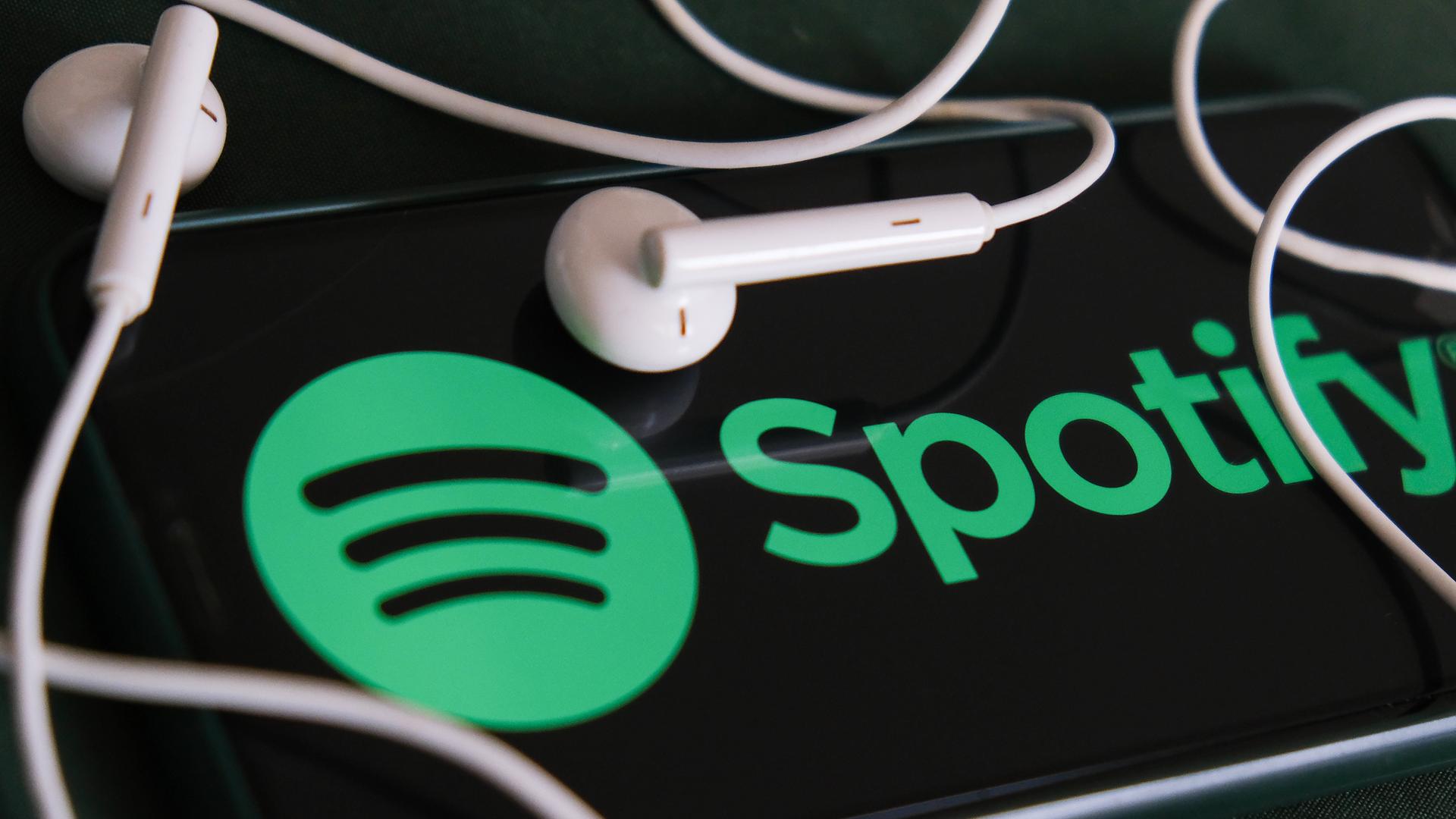 Das Foto zeigt ein Handy-Bildschirm, auf dem das Spotify in grüner Schrift angezeigt wird, und zwei Kopfhörer-Kapseln.