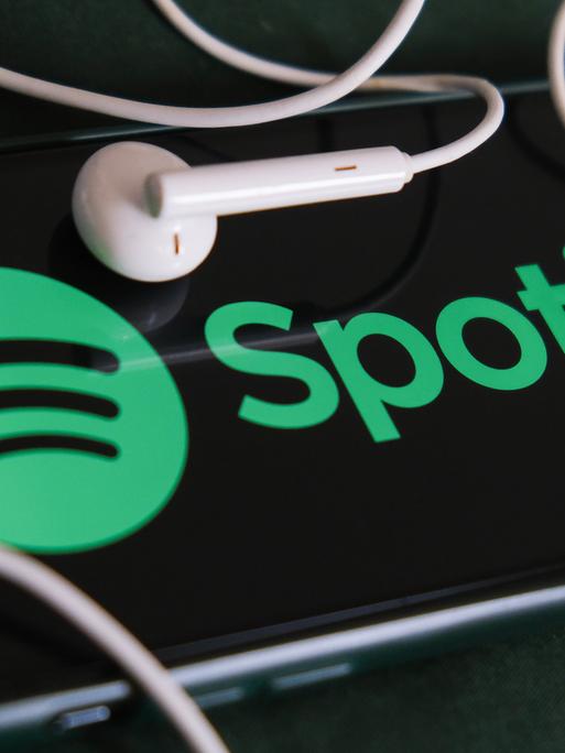 Das Foto zeigt ein Handy-Bildschirm, auf dem das Spotify in grüner Schrift angezeigt wird, und zwei Kopfhörer-Kapseln.