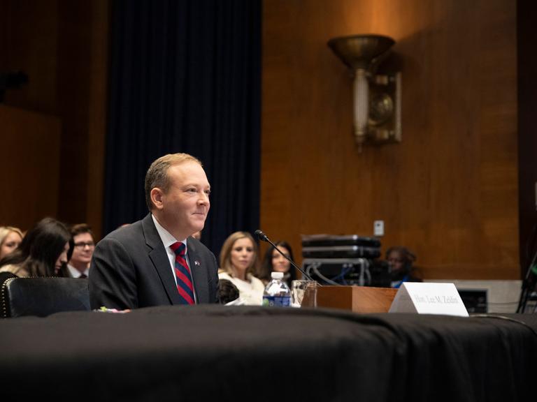 Der US-Rebublikaner Lee Zeldin sitzt an einem Pult. Hinter ihm sind mehrere Personen. 