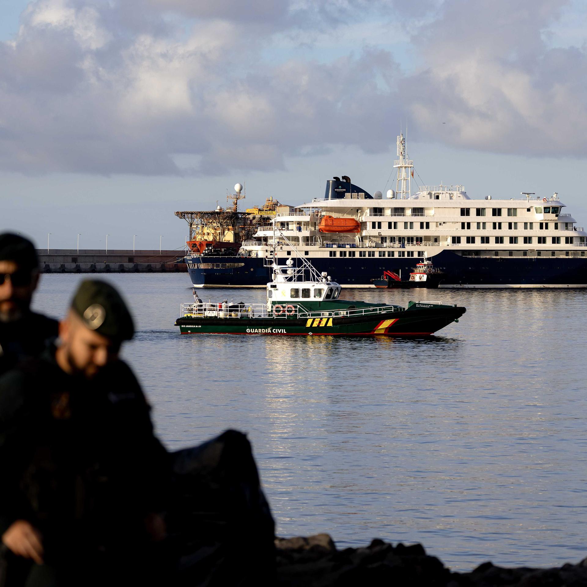 Das Kreuzfahrtschiff "Hondius" liegt im Wasser vor einer Küste, daneben ist ein Schiff der Küstenwache.