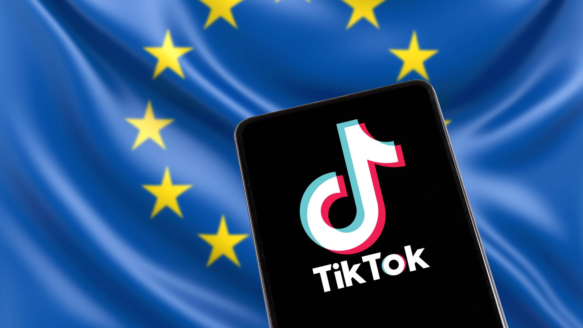 Das Foto zeigt ein Smartphone, auf dem das Logo der App TikTok angezeigt wird. Im Hintergrund ist eine Europa-Fahne zu sehen.